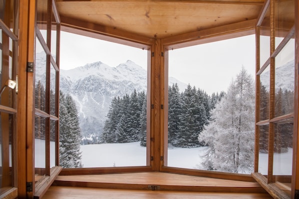 A room with a large wooden-framed window offering a view of snow-covered mountains and trees.