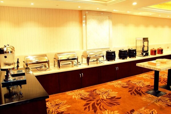 Área de buffet de hotel com diversos pontos de comida, uma máquina de café e uma pequena mesa com um bolo.