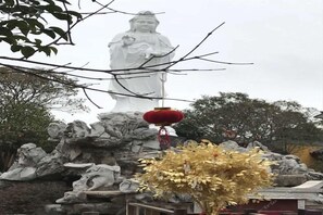 Une grande statue blanche, devant laquelle se trouve une lanterne rouge, le tout entouré de rochers et de feuillage.