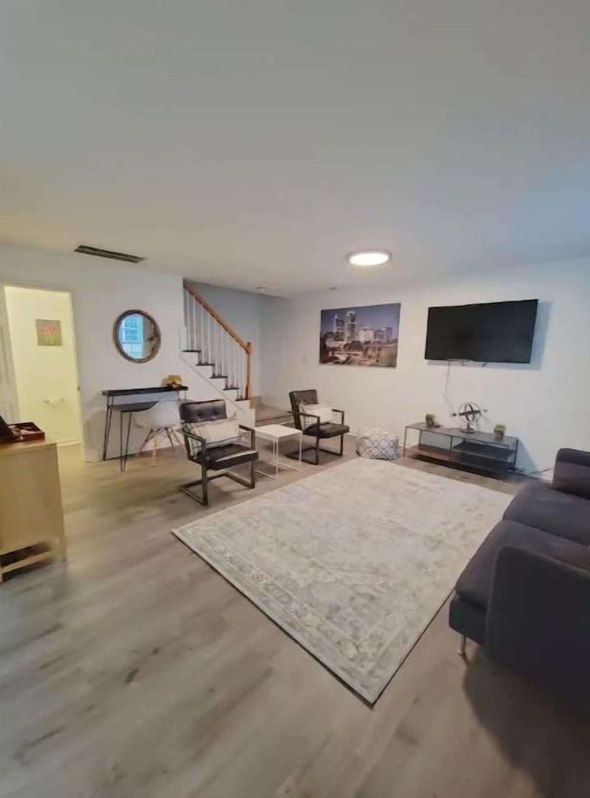 A living room with a staircase, a flat-screen TV mounted on the wall, and a large rug.