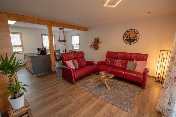 Un salon avec un canapé rouge, une table basse, un plancher en bois, une cuisine à l’arrière-plan et une horloge au mur.