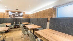 Ein modernes Restaurant mit Holztischen und grauen gepolsterten Sitzbänken, einem großen Fenster und einem Poster mit Berglandschaft an der Wand.