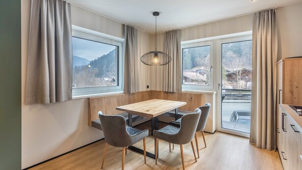 Ein Essbereich mit einem Holztisch, vier Stühlen, großen Fenstern mit Vorhängen und einer hängenden Lampe.
