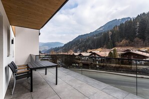 Un balcon avec une table et des chaises, donnant sur une route et des montagnes boisées.