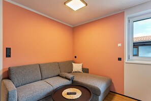 Una habitación con un sofá gris, una pequeña mesa redonda con un control remoto y una ventana que ofrece vistas a un edificio.