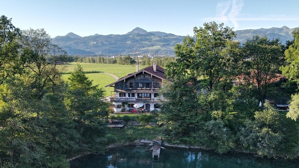 A large wooden house with a balcony, surrounded by lush greenery and a lake, with mountains in the background. - Ferienwohnung bis 4 Personen (Kiefersfelden)