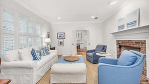 Ein Wohnzimmer mit einem weißen Sofa, einem blauen Sessel und einem Hocker, einem Kamin und Blick zur Küche.