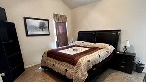 Un dormitorio con cama, ventilador de techo, mesita de noche, cubo de basura, un cuadro en la pared y una puerta.