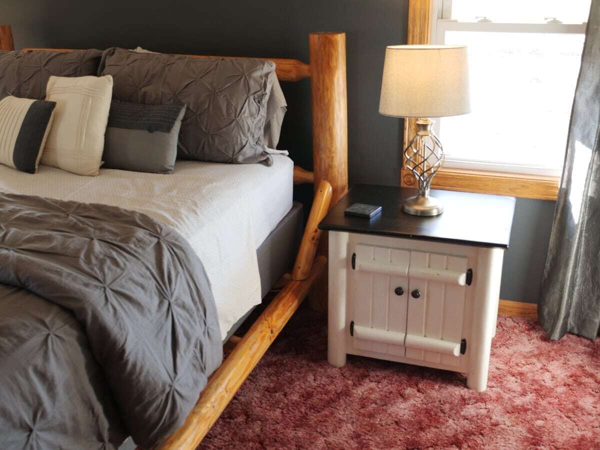 Una cama con edredón gris, una mesita de noche con lámpara y una ventana con cortinas.