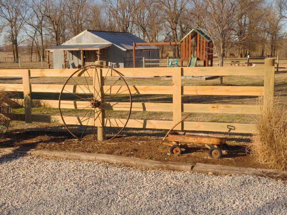 Un décor extérieur rustique agrémenté d’une clôture en bois, d’une grande roue et d’un chariot en bois.