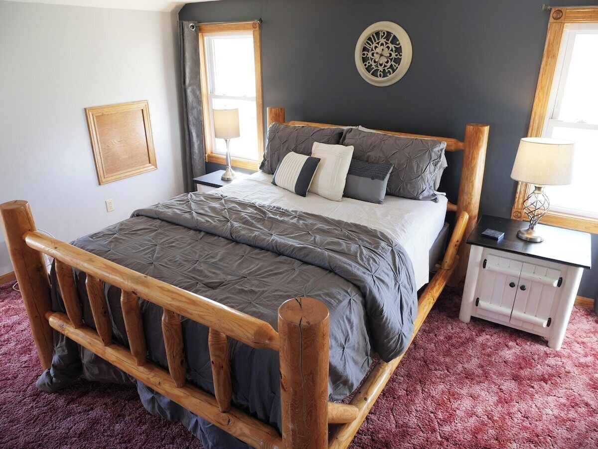 Una cama con estructura de madera, colchón acolchado grisáceo y almohadas. Hay una mesita de noche blanca con una lámpara, un reloj de pared y un cuadro enmarcado.