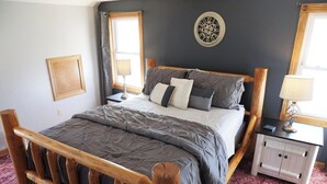 A bed with a wooden frame, a grey quilted mattress, and pillows. There is a white nightstand with a lamp, a wall clock, and a framed picture.