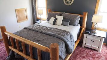 Una cama con estructura de madera, colchón acolchado grisáceo y almohadas. Hay una mesita de noche blanca con una lámpara, un reloj de pared y un cuadro enmarcado.