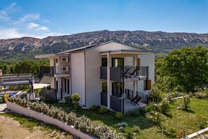 Una casa moderna de dos pisos con balcón, rodeada por un jardín y montañas al fondo.