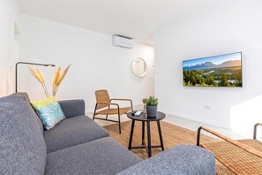 A modern living room with a grey sofa, a round mirror, a wicker chair, a small table with a potted plant, and a wall-mounted TV displaying a scenic landscape. - Pool Apartment Sapore Di Mare 3, Baška, Croatia (Baška)