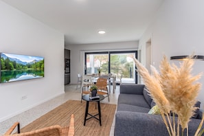 Un moderno salón con un televisor de pantalla plana, un sofá gris, una zona de comedor con mesa y sillas, y vistas a un paisaje montañoso a través de grandes ventanales.