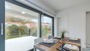 A dining area with a table set for two, a potted plant, and a view of a residential area through large windows.