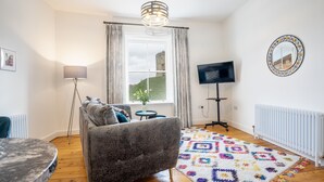 Ein Wohnzimmer mit grauem Sofa, rundem Couchtisch, farbenfrohem Teppich, Fernseher auf einem Lowboard und einem Fenster mit Vorhängen.