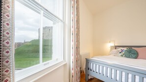 Ein Schlafzimmer mit Holzboden, einem weißen Bett mit blauer Kopfteil und einem Fenster mit gemusterten Vorhängen, mit Blick auf eine grüne Landschaft.