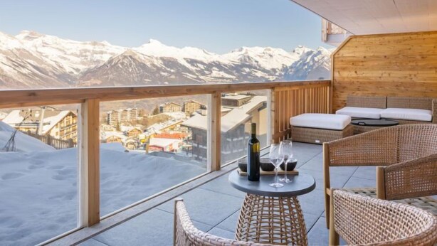 A balcony with a view of snow-covered mountains, wicker furniture, and a bottle of wine. - Les Terrasses de Nendaz D1 by Interhome (Nendaz)