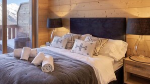 A bedroom with a wooden wall, a bed with white bedding, two bedside lamps, and a view of mountains through a sliding door. - Les Terrasses de Nendaz D1 by Interhome (Nendaz)