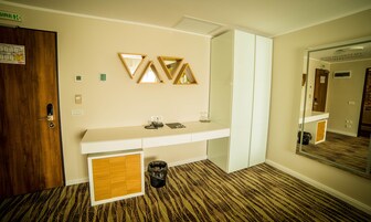 A hotel room with a wooden door, a white vanity with a mirror, a small trash bin, and a patterned carpet.