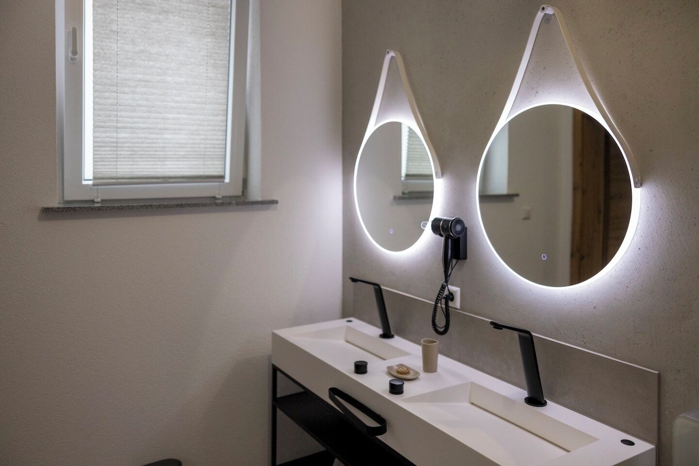 A bathroom with a white vanity, two mirrors with circular light fixtures, a telephone, and a window with blinds.