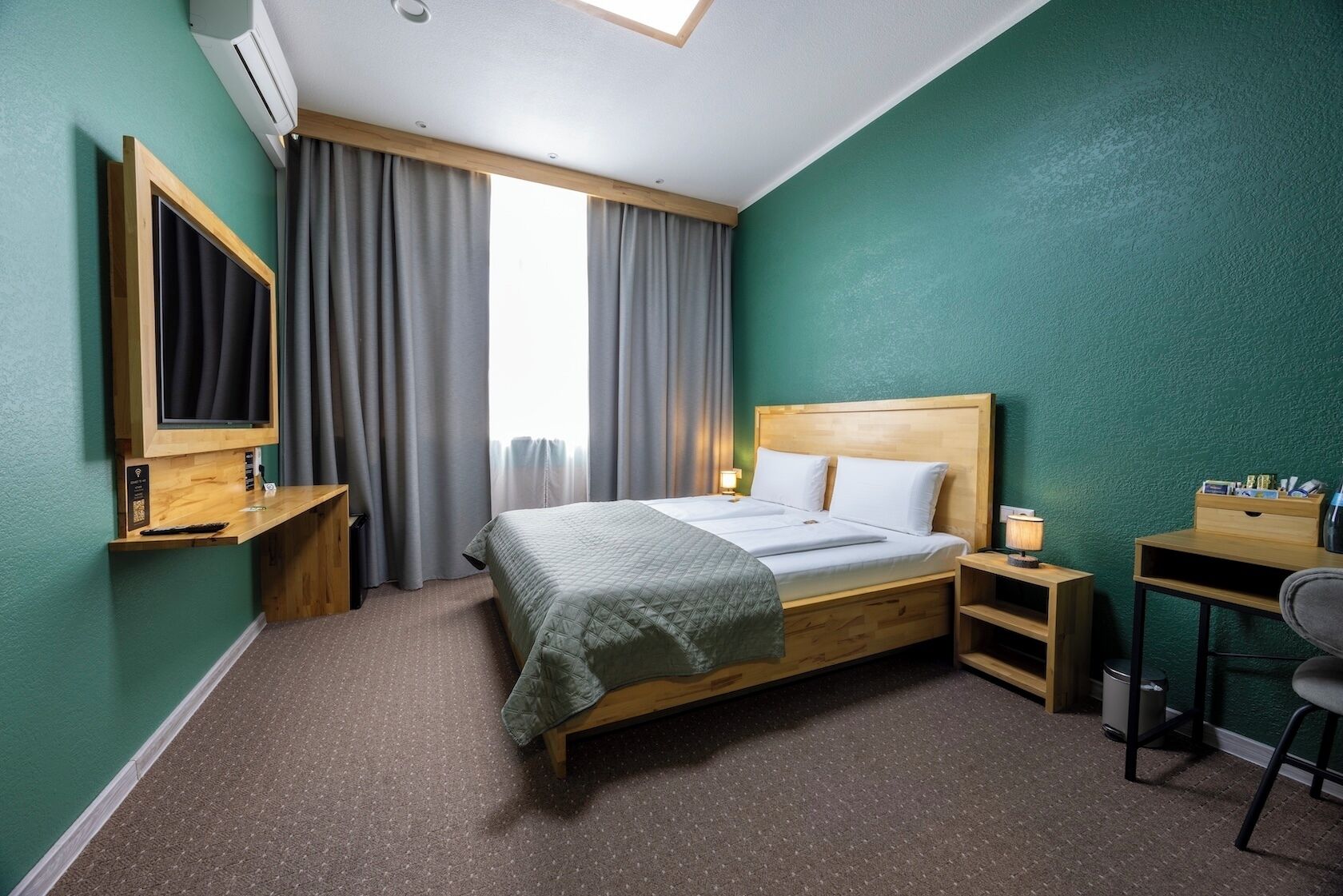 Quarto de hotel com cama, escrivaninha, cadeira e televisão fixada na parede.