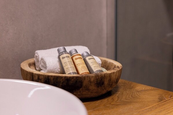 A wooden bowl containing three bottles of Touch Naturals shampoo and body lotion, placed on a wooden surface next to a white sink.