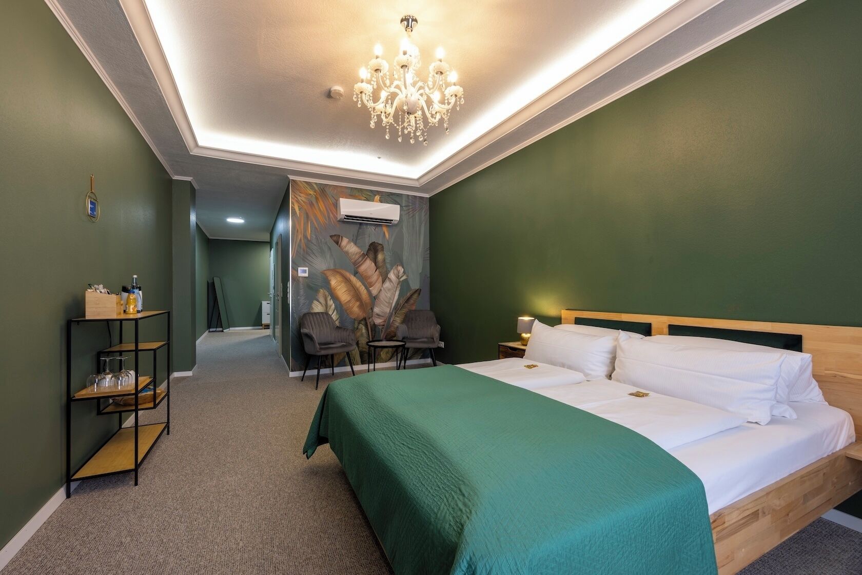 A hotel room with a bed, a chandelier, a wall-mounted air conditioner, a small table with a lamp, and a chair.