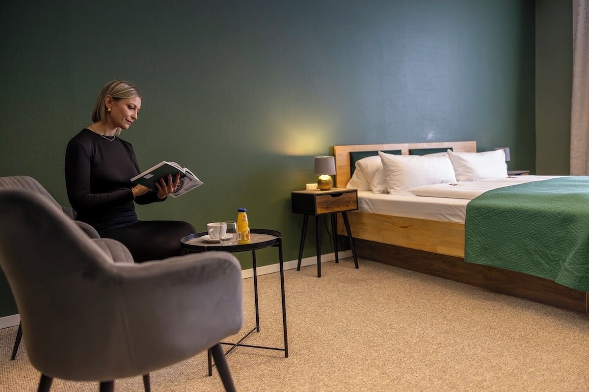 A hotel room with a bed, a bedside table with a lamp, a chair, and a small table with a cup and a bottle.