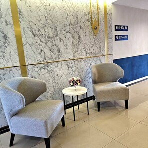 A hotel lobby with two armchairs, a small round table with a vase of flowers, marble walls, and directional signs with phone numbers. - Le Srivaree Suvaranabhumi Airport Hotel (Bang Phli)