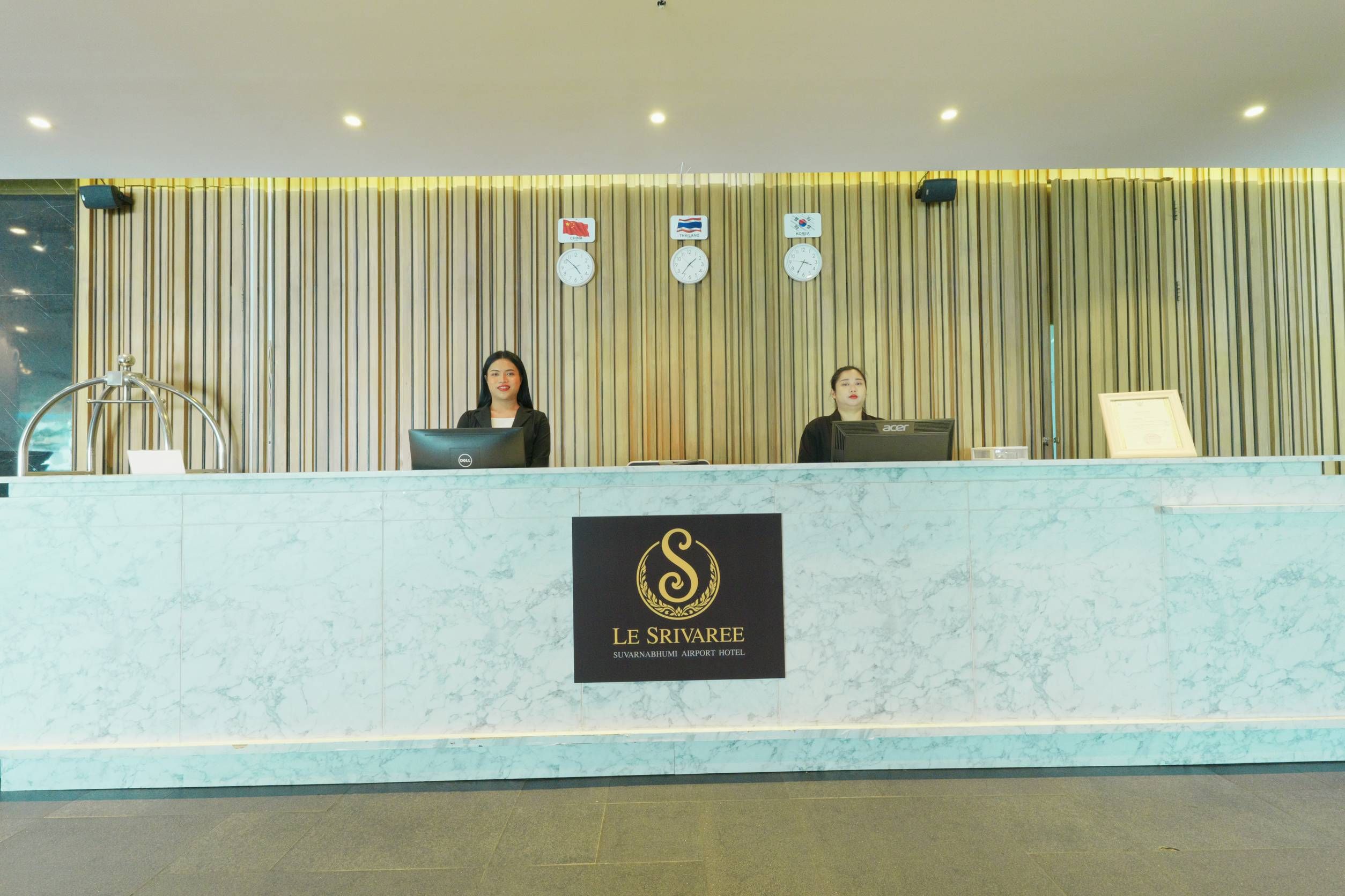 A reception desk with a marble top and a logo for Le Sirvaee Suvarnabhumi Airport Hotel. There are clocks displaying time from different countries and a computer on the desk.