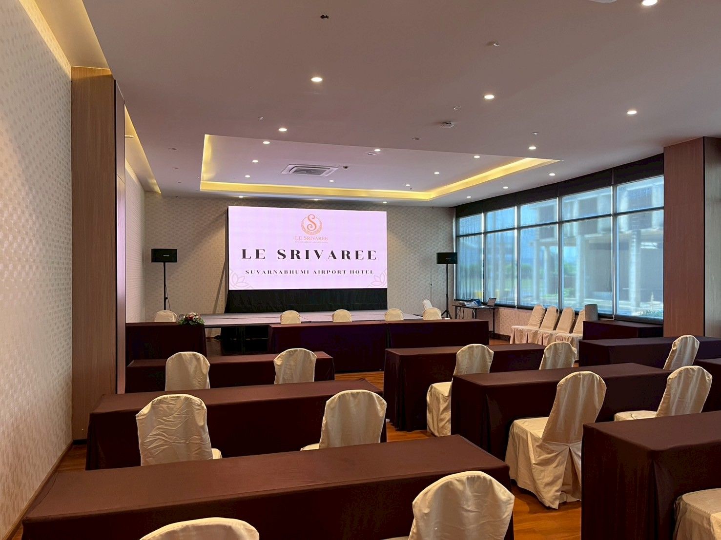 A conference room with a stage, rows of tables, and chairs, and a large screen displaying 'LE SRIVAREE'.
