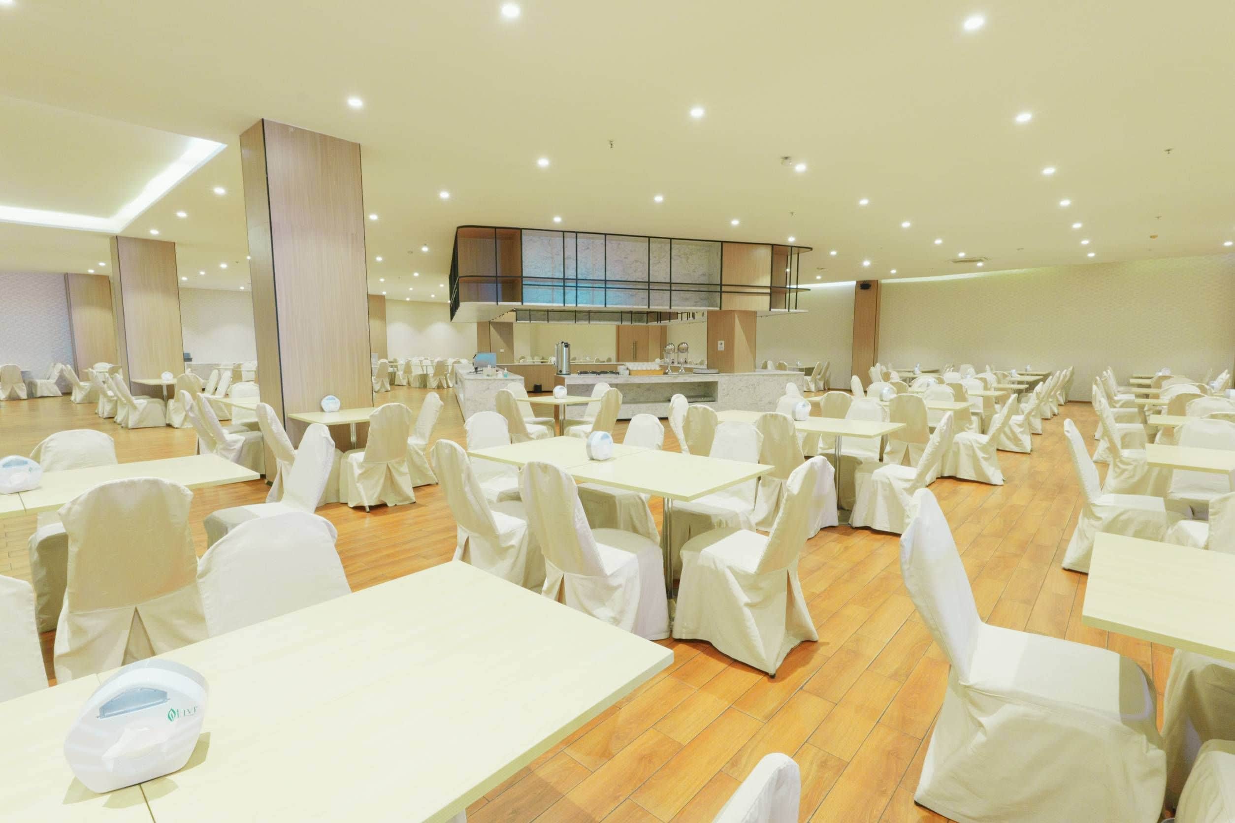 A spacious banquet hall with multiple tables and chairs, a central kitchen area, and a high ceiling with recessed lighting.