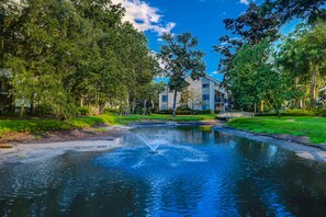 Surrounded by lush greenery and mature trees, this lakeside setting features a tranquil fountain and a modern building with balconies and large windows.