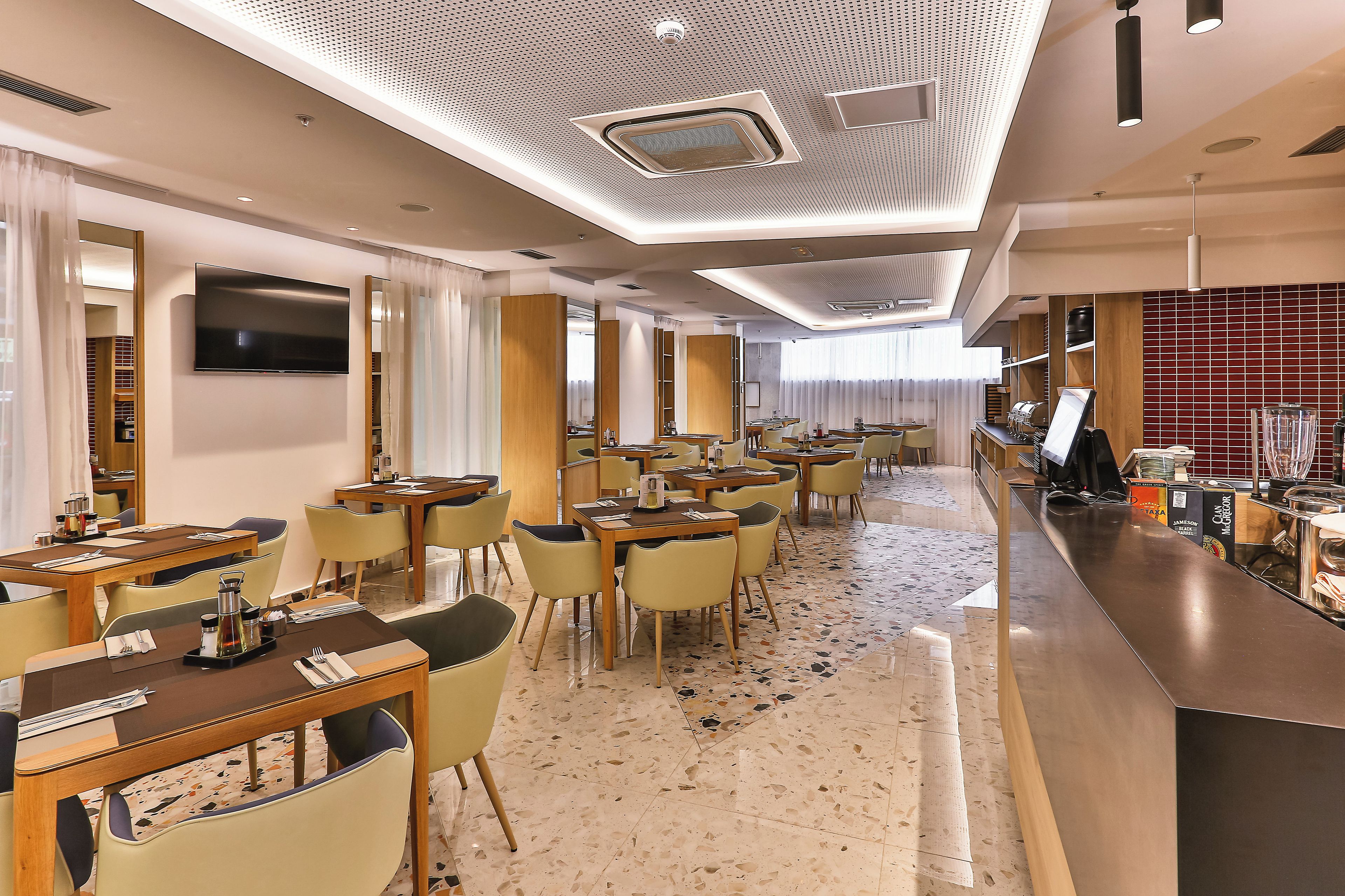 Un comedor moderno de hotel con varias mesas y sillas, un televisor de pantalla plana en la pared, y una zona de bar con una máquina de café.