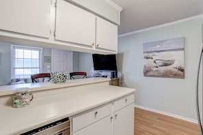 A kitchen with white cabinets, a countertop, a window with blinds, a television, and a painting of a beach scene.