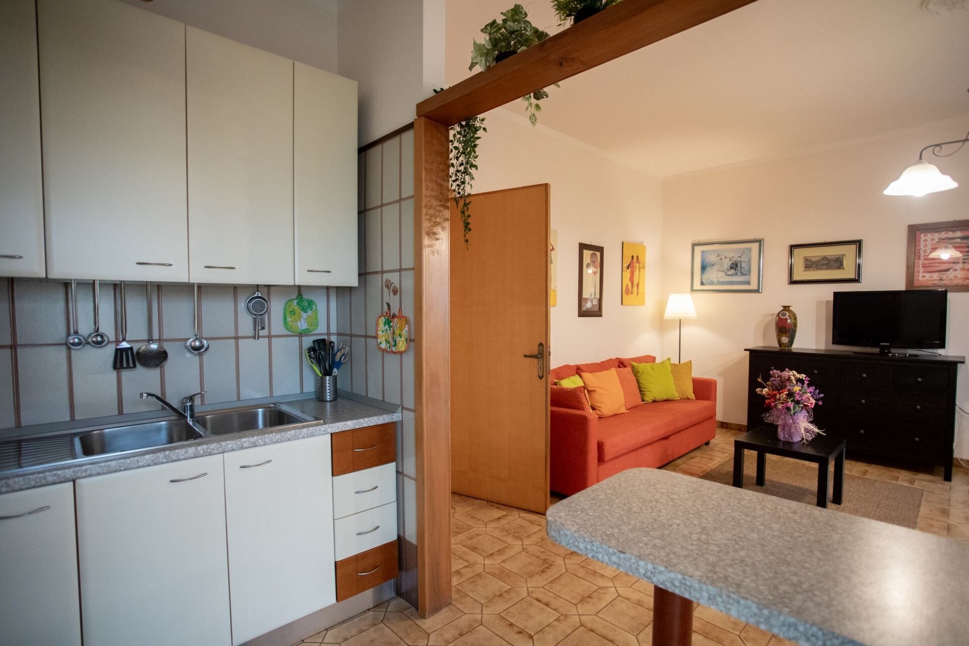Una cocina con armarios blancos, un lavamanos y utensilios colgando. Hay un sofá rojo, una mesita y un televisor en la sala.