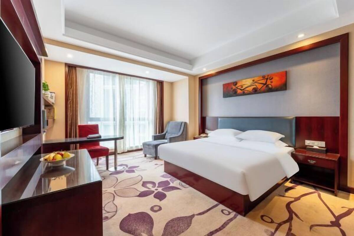 Une chambre d’hôtel avec un lit, un bureau sur lequel se trouve un panier de fruits, une chaise, un téléviseur et un tableau accroché au mur.