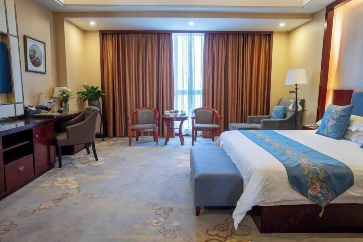 Une chambre d’hôtel avec un lit, deux chaises, un bureau, une lampe et une fenêtre avec des rideaux.
