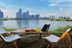 A lakeside view with a city skyline in the background, a picnic table with food and drinks, and two lawn chairs facing the water. - Hengsheng International Hotel (Yiwu)