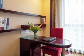 A hotel room with a glass table, a red chair, a vase with flowers, and a bookshelf with books and a box.