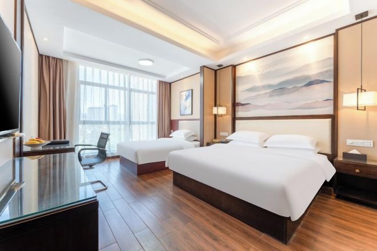 A hotel room with two beds, a desk, a TV, and a large window.