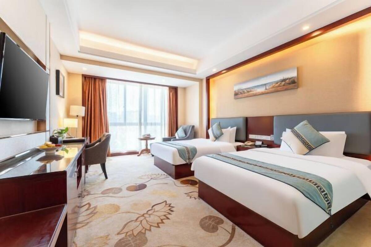 Une chambre d’hôtel avec deux lits, un bureau avec une télévision, une petite table et une fenêtre avec des rideaux.