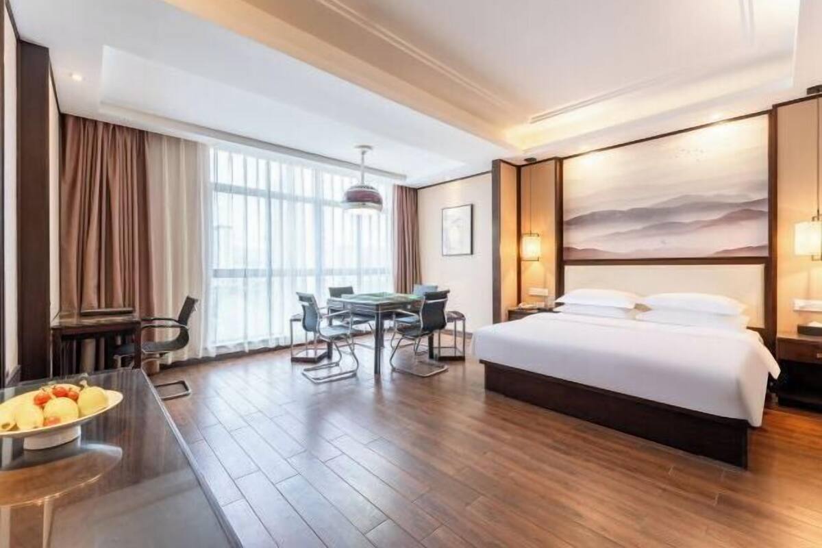 Une chambre d’hôtel avec un grand lit, un bureau avec une chaise, une table à manger et une grande fenêtre avec des rideaux.