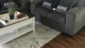 Ein Wohnzimmer mit grauem Sofa, zwei Kissen mit dem Aufdruck 'HOME', einem kleinen Blumentopf, einer Fernbedienung und einem weißen Couchtisch.