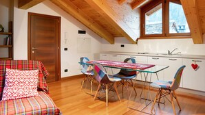 A cozy room with a wooden ceiling, a dining table with colorful chairs, a sofa with plaid bedding, and a window overlooking a snowy landscape.