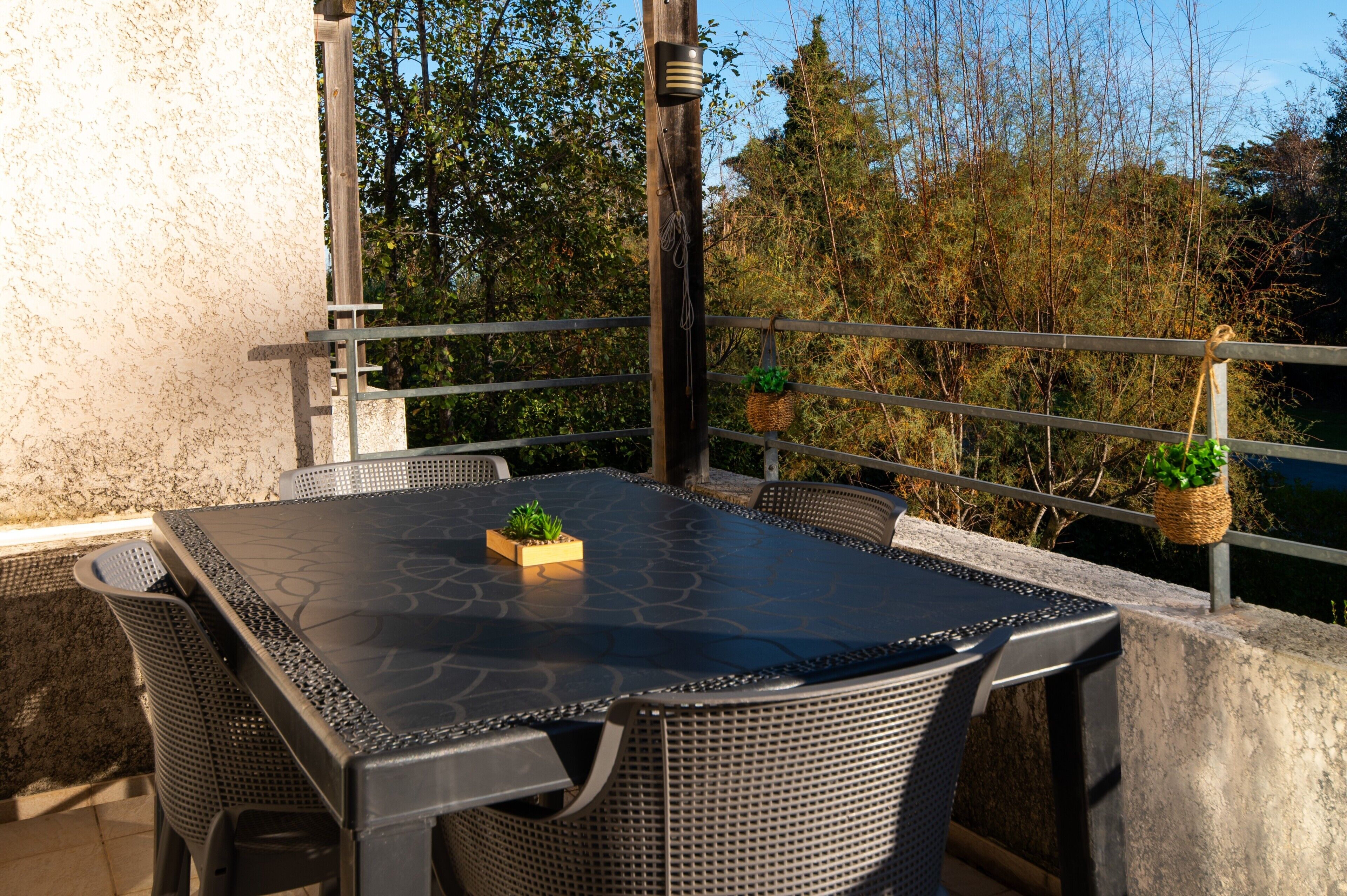 Une terrasse extérieure couverte, dotée d’une table et de chaises, et entourée d’une rambarde et de verdure.