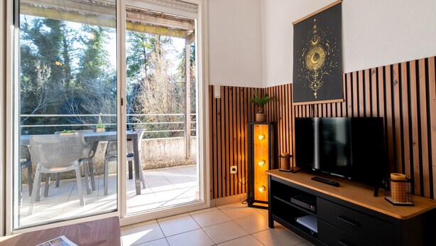 A modern living room with a TV, wooden panel wall, sliding glass door to a balcony, and a magazine on a table. - Holiday Apartment "Poggio Mezzana" with Sea View, Shared Pool & A/C (Santa-Lucia-di-Moriani)